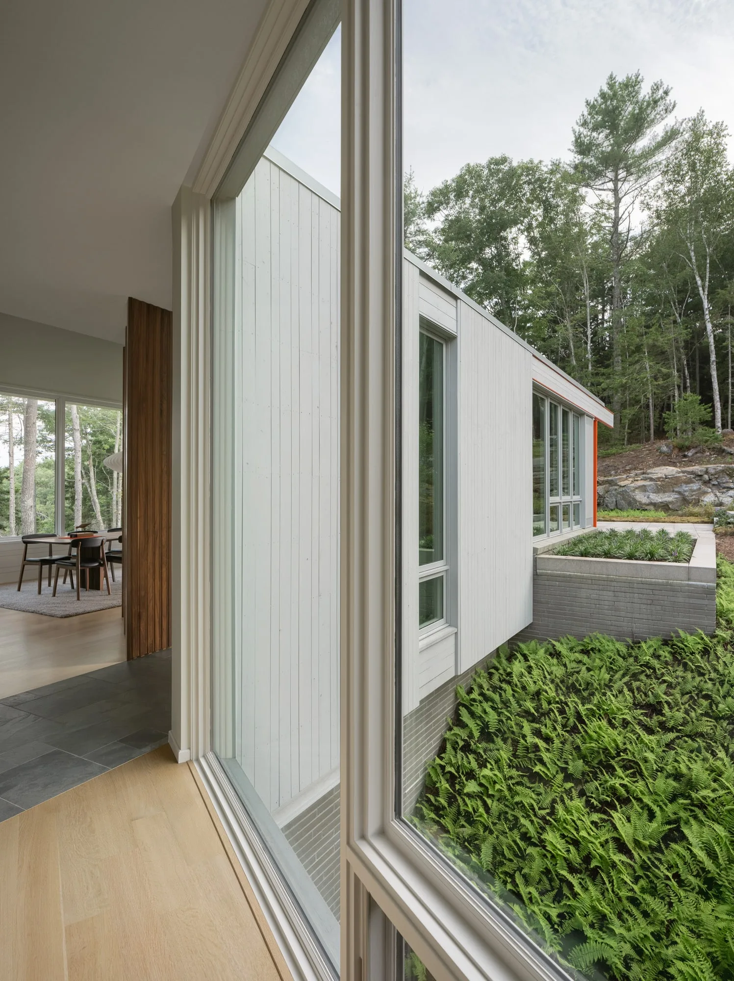 Side exterior view of a high-end modern home in Maine, with large glass windows overlooking lush greenery and a serene wooded landscape