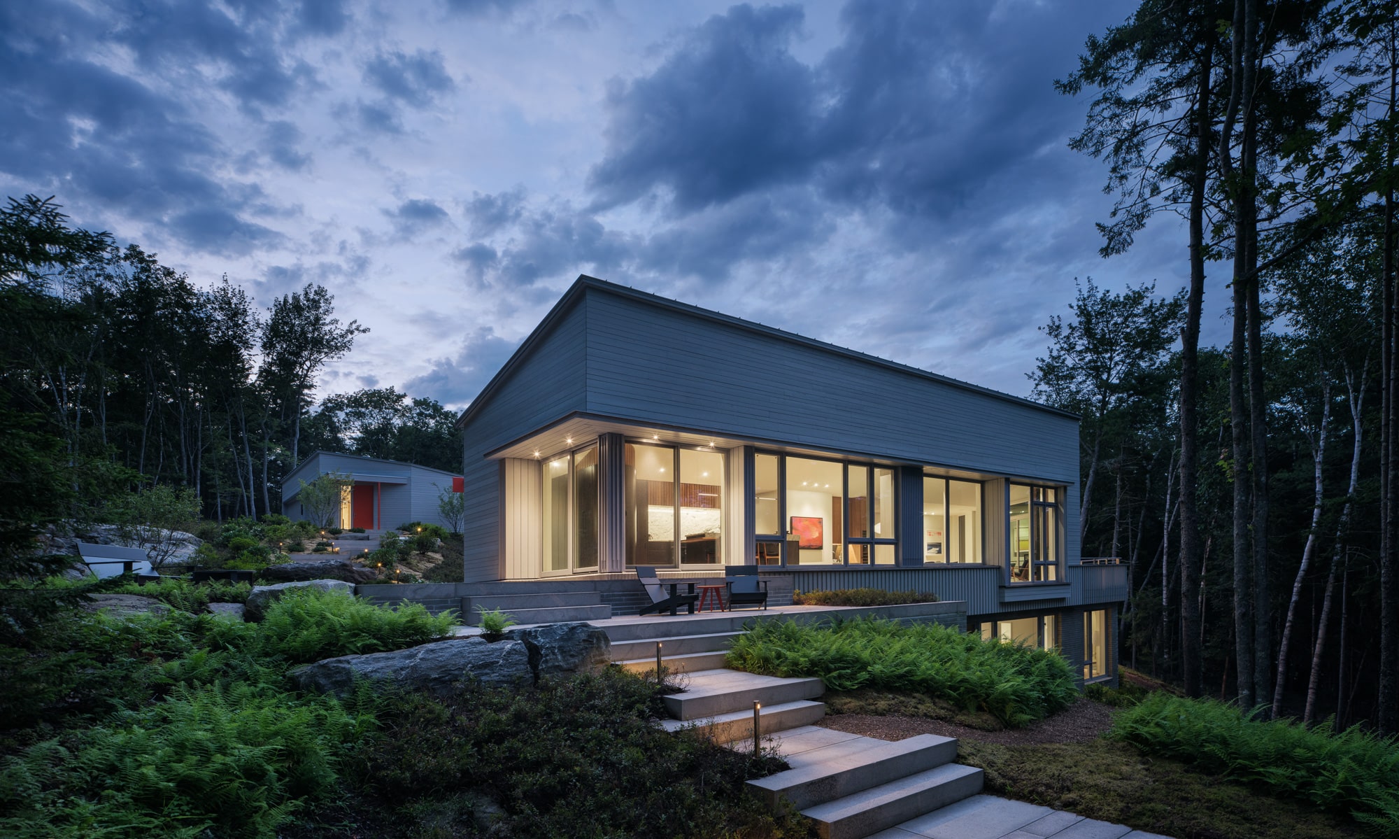 Green Haven’s modern exterior at dusk, with warm interior lighting glowing through tall windows and lush greenery surrounding the home