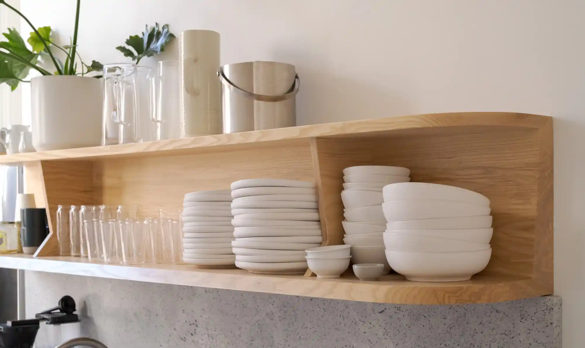 Closeup of light wood shelves in the office kitchen with off-white plates, bowls, and glass cups, showcasing a clean, modern design with natural elements.