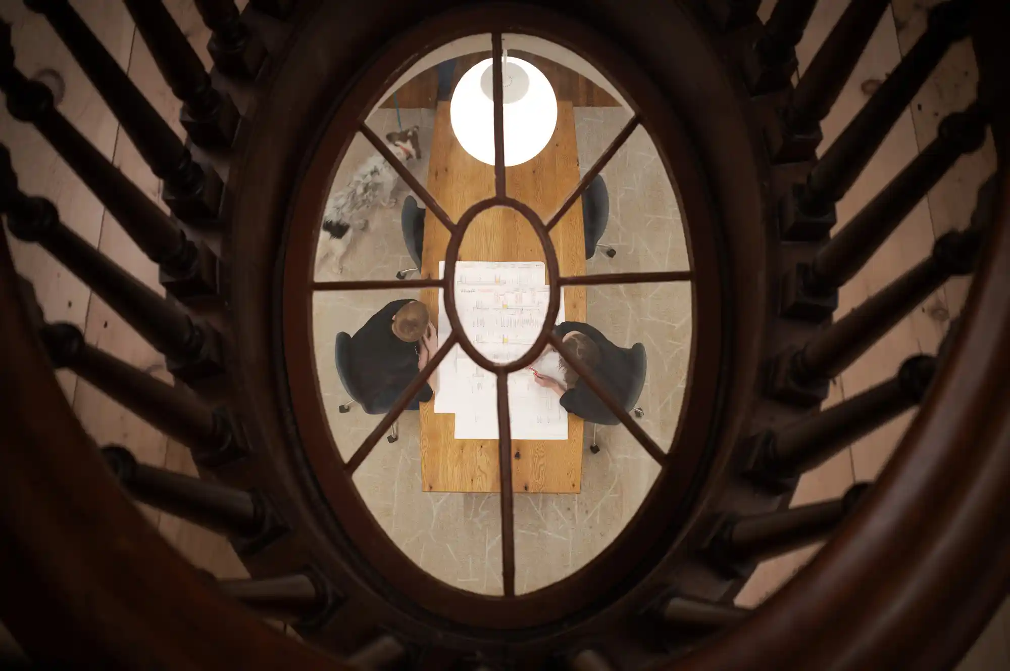 View from an oval window on the second floor of the Safford House, looking down at two people reviewing architectural designs at a rectangular table.