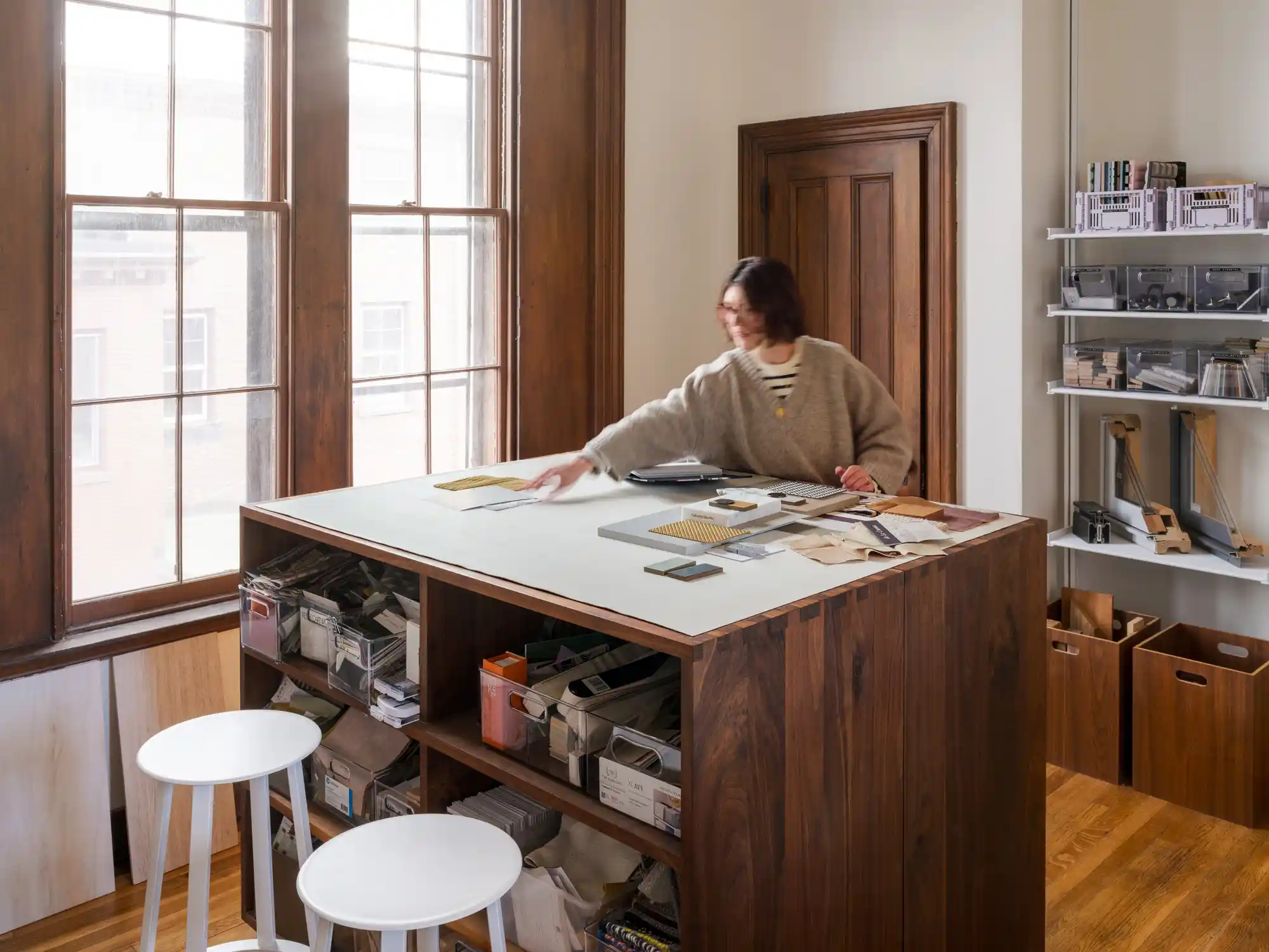 A woman standing at a dark wood island in a spacious office, with historic wood-framed windows and open shelving filled with design supplies at Woodhull's workspace.