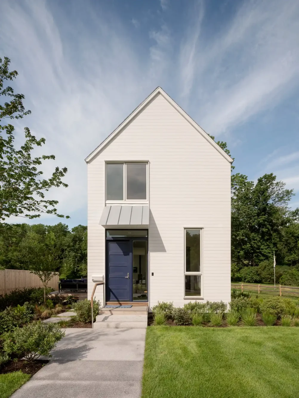 Front view of a contemporary 16-foot-wide custom home in Cape Elizabeth, Maine, designed by Woodhull. Features white cedar siding, a blue door, manicured landscaping, and a modern architectural style.