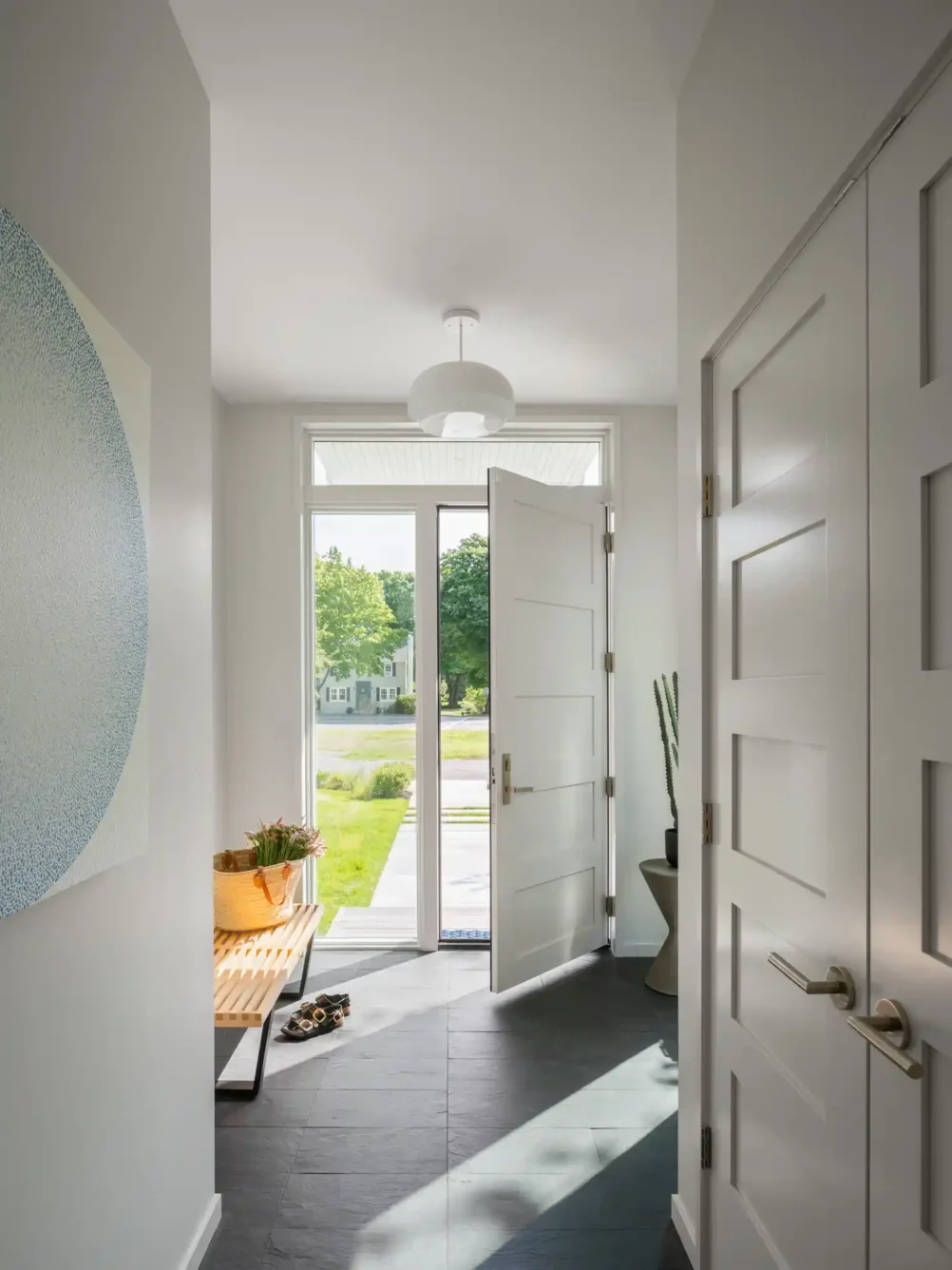 Bright and airy entryway of a custom home in Cape Elizabeth, Maine, designed by Woodhull. Features slate flooring, a white front door, and natural light highlighting minimalist interior design.