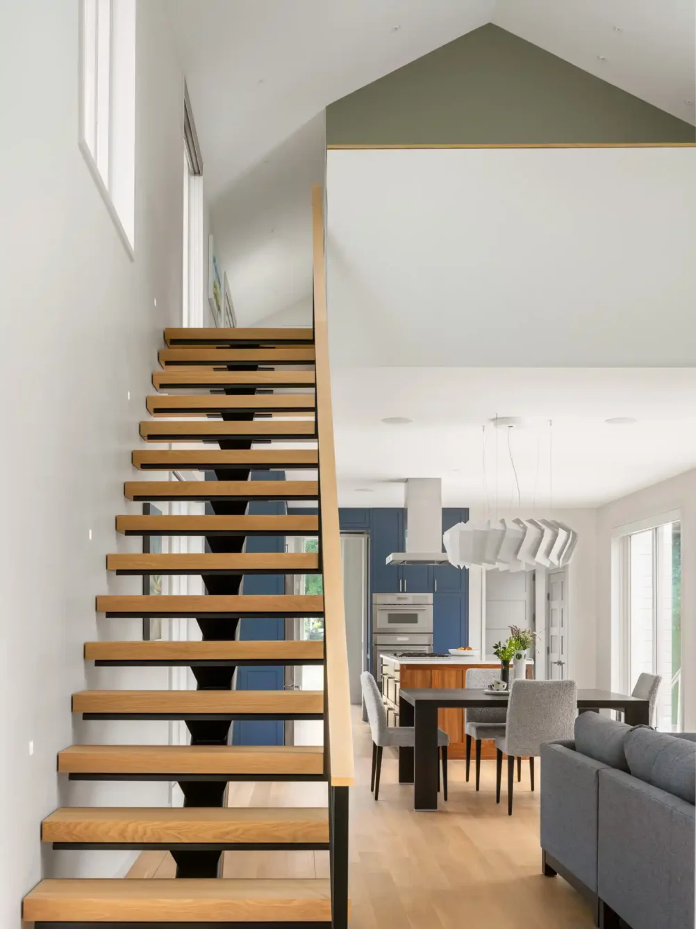 View of floating wood stairs leading to a lofted area in a custom home designed by Woodhull in Cape Elizabeth, Maine. A glimpse of the first floor features a gray sofa, dark wood dining table with gray fabric chairs, and a modern kitchen with blue cabinets in the background.