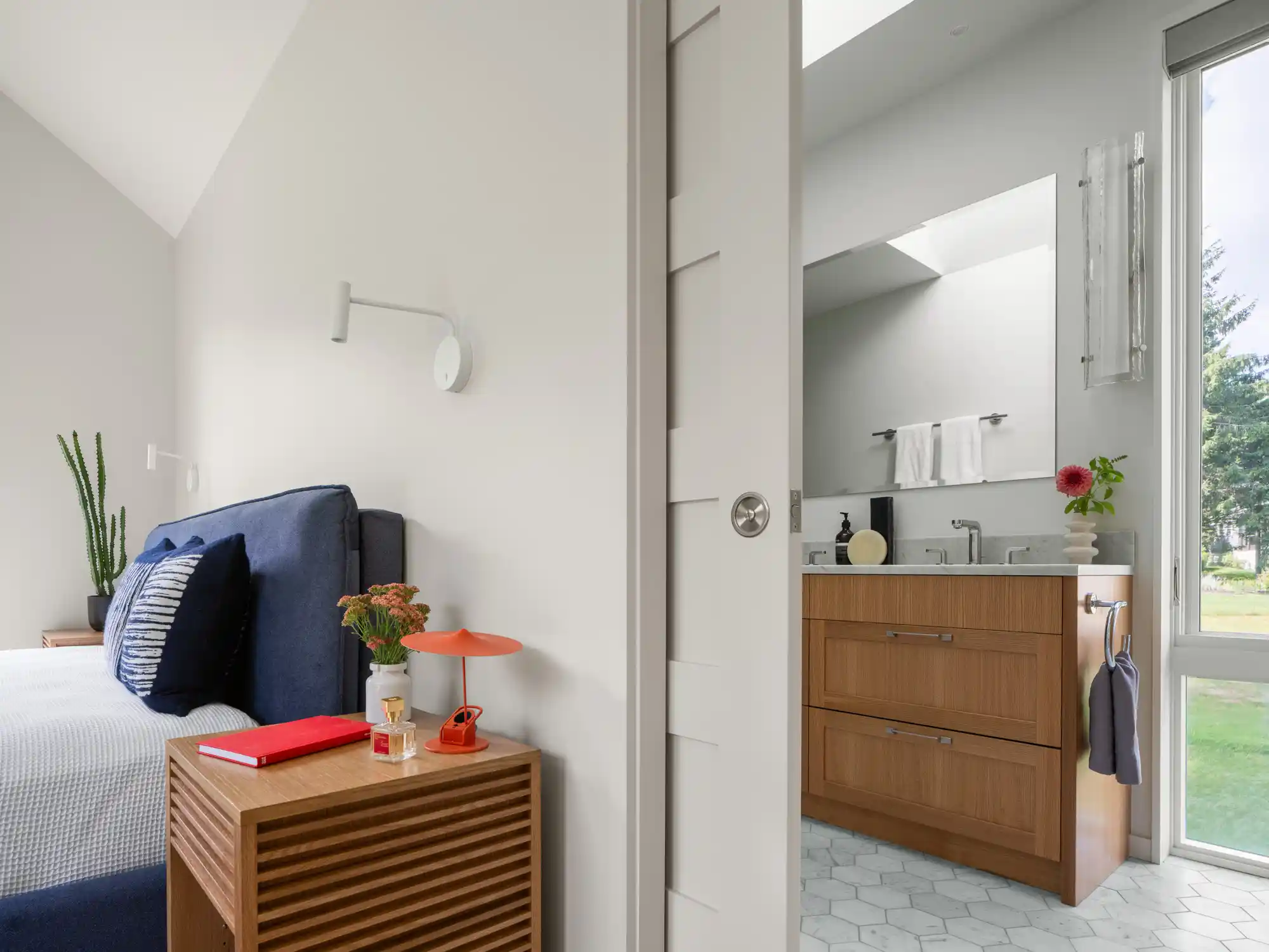 Split view of a modern bedroom and bathroom in a custom home designed by Woodhull in Cape Elizabeth, Maine. The bedroom features a blue fabric headboard and wood nightstand, while the bathroom showcases a wood vanity, a pocket door, and a large floor-to-ceiling window that fills both spaces with natural light.