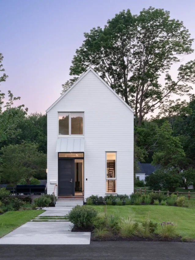 Exterior of a narrow, modern home in Cape Elizabeth, Maine, designed by Woodhull, showcasing white cedar siding, a bold blue front door, and a landscaped pathway.