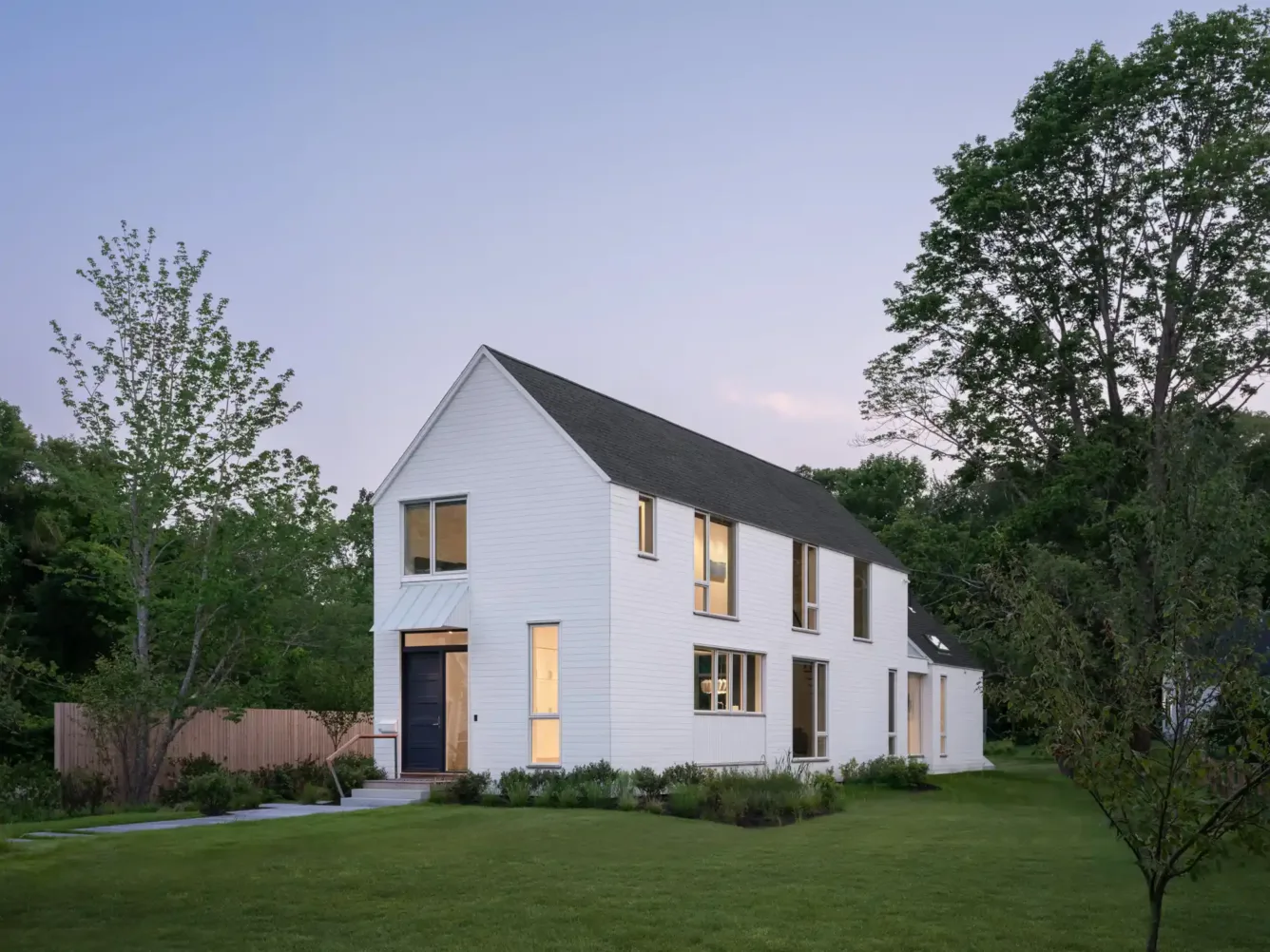 Front view of a contemporary 16-foot-wide custom home in Cape Elizabeth, Maine, designed by Woodhull. Features white cedar siding, a blue door, manicured landscaping, and a modern architectural style.