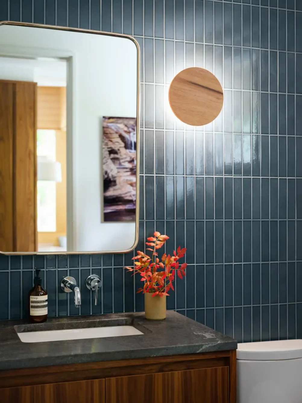 A contemporary bathroom featuring a dark wood vanity with a black stone countertop, chrome fixtures, and vertically laid dark blue tiles. The mirror has a brass-trimmed frame that adds a touch of warmth to the modern design.