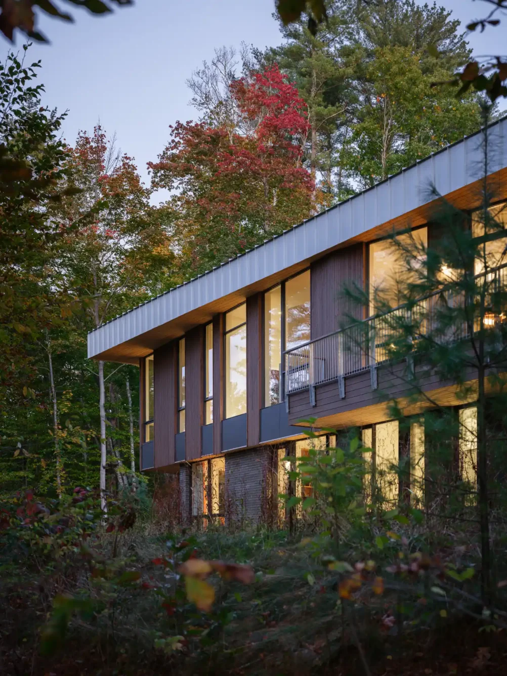 A rear view of a secluded modern home in Maine, featuring a wraparound balcony and surrounded by lush woodland. The home is designed to blend into its environment while offering expansive views of the natural landscape.