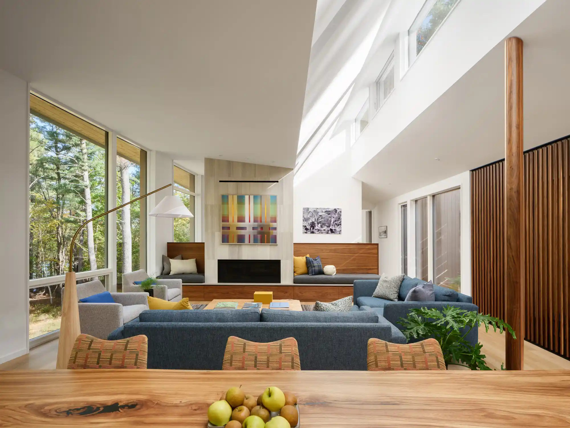 A view from the kitchen into the living room, featuring a denim blue sofa, a natural wood dining table, and a black fireplace. Floor-to-ceiling windows bring in natural light and frame the lush outdoor scenery.