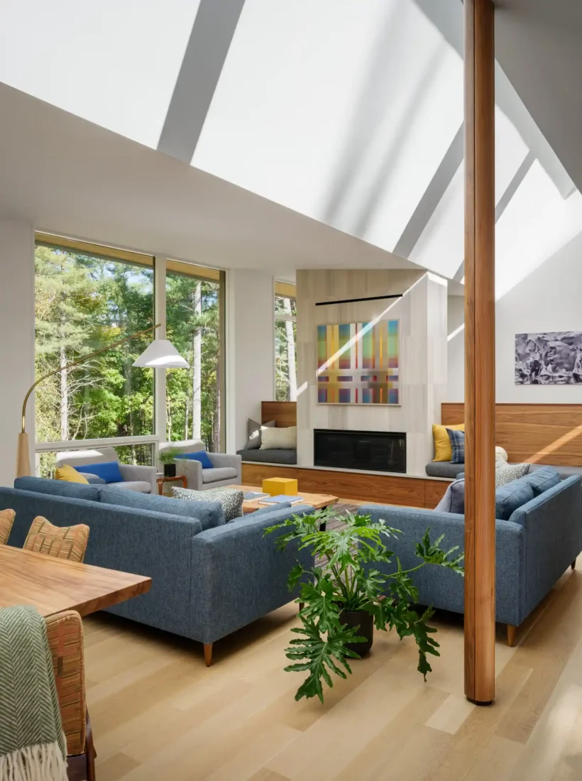 An open living area featuring a denim blue sofa, yellow accent pillows, and expansive windows with views of the surrounding forest. The mix of modern and natural materials creates a balanced and inviting space.