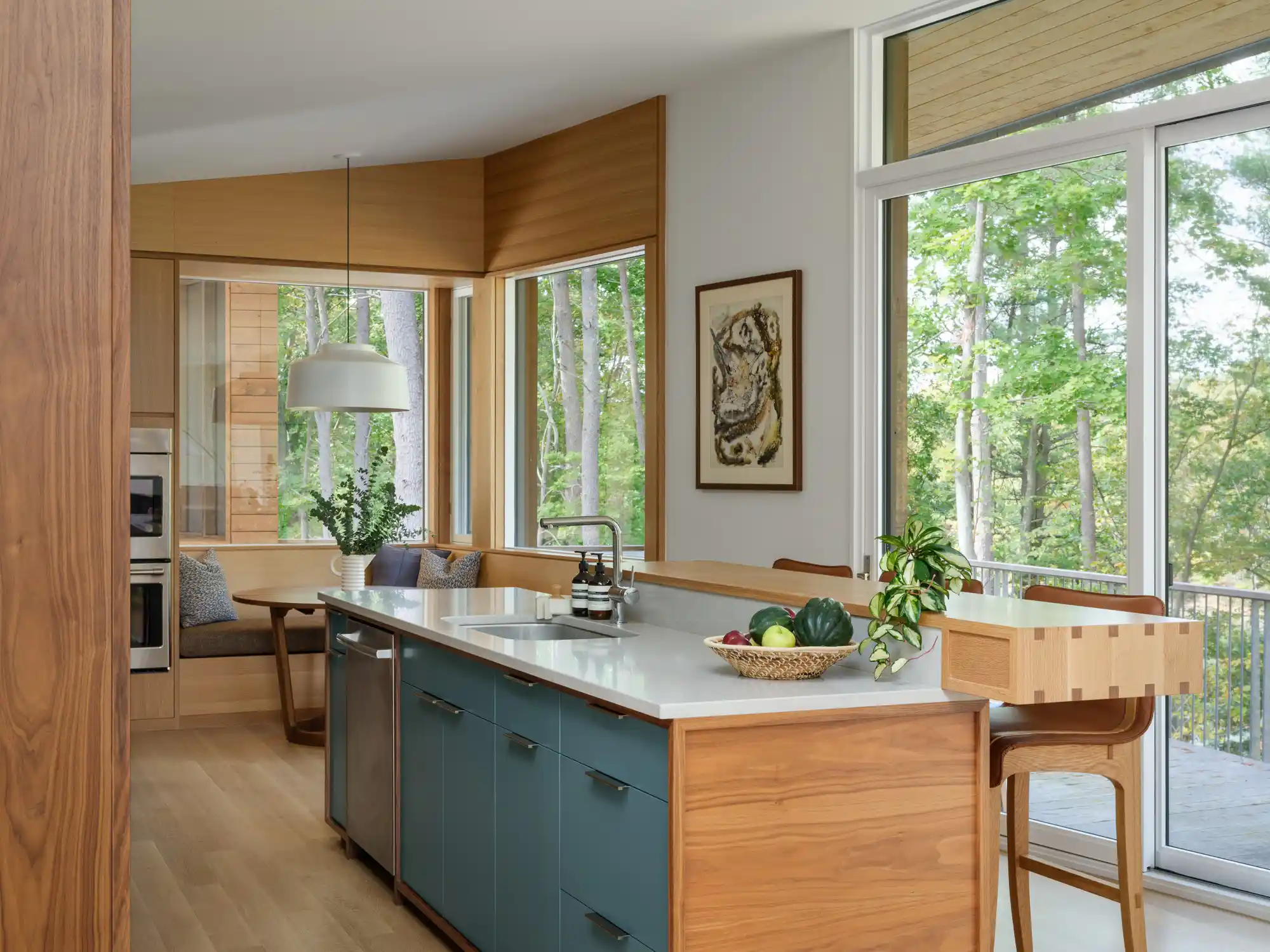 A modern kitchen with a wood-accented island, light blue cabinetry, a stainless steel dishwasher, and a view of the forest through sliding glass doors. The combination of natural wood tones and soft blue hues adds warmth to the contemporary design.