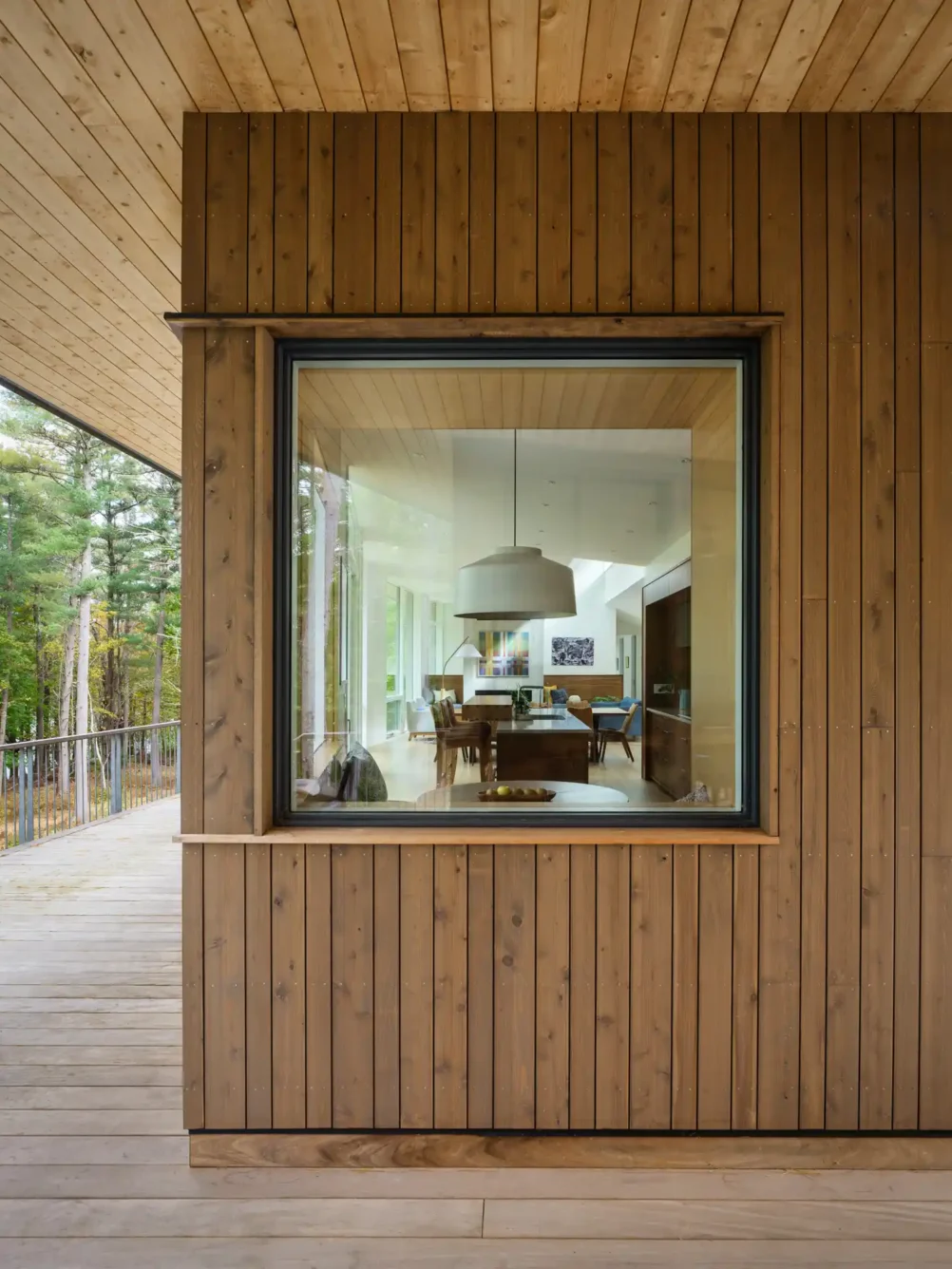 A perspective from a wood-paneled balcony, looking through a large square window into the kitchen and living room. The open-concept design allows for uninterrupted sightlines, enhancing the connection between indoor and outdoor spaces.