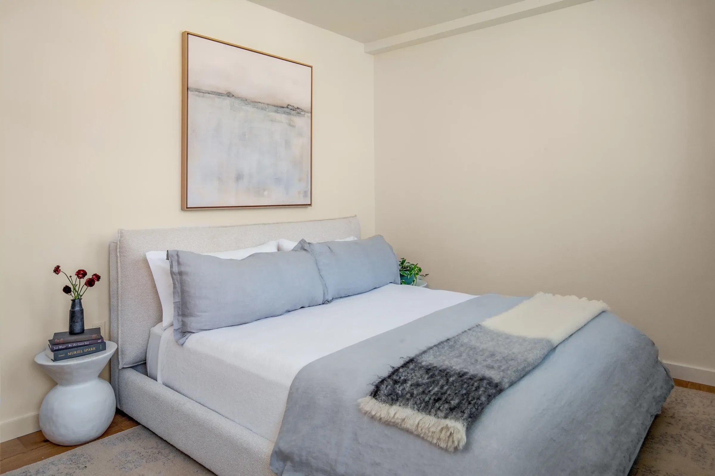 Tranquil bedroom at the Weldon Hotel, constructed by Woodhull, featuring a low-profile upholstered bed with soft blue-gray bedding, abstract wall art, and a sculptural side table with books and flowers