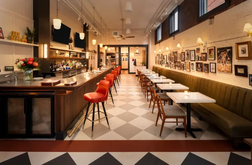 Interior of Benny’s Restaurant featuring a long olive green banquette, bistro-style chairs, cherry red bar stools, a fully stocked bar, and checkered flooring