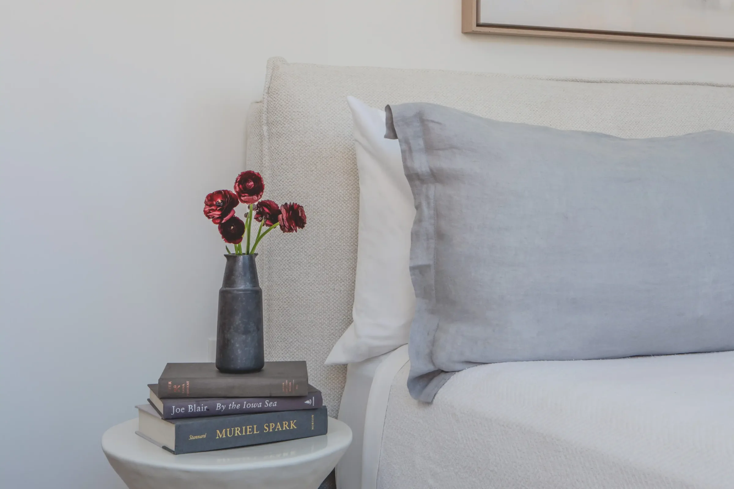 Close-up of bedside detail at the Weldon Hotel, constructed by Woodhull, showing a soft upholstered bed, linen pillow, and a sculptural side table with stacked books and a vase of red flowers
