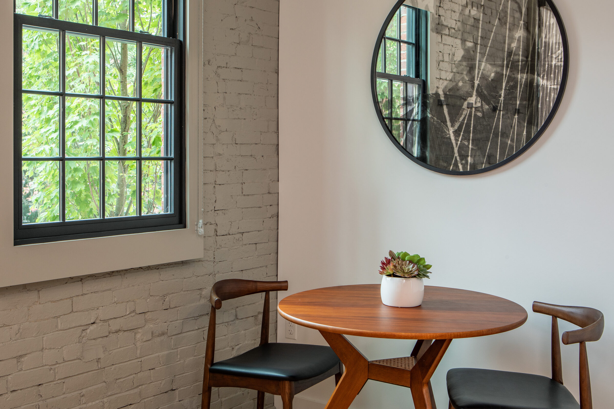 Cozy dining nook at the Weldon Hotel, constructed by Woodhull, featuring a round wood table with two chairs, a potted succulent centerpiece, brick walls, and a round mirror reflecting industrial details