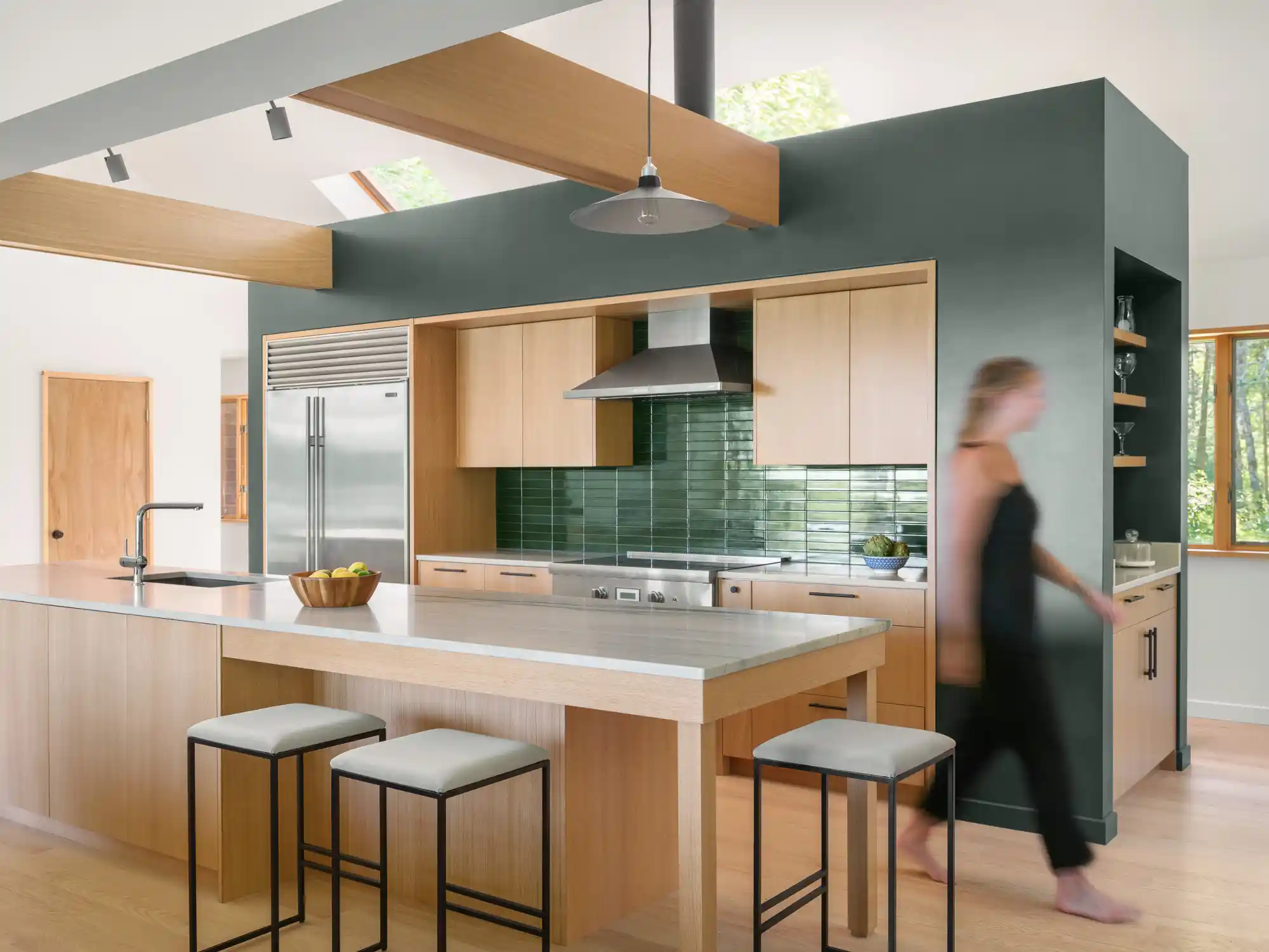 Contemporary kitchen with light wood cabinetry, green walls, stainless appliances, and person walking through