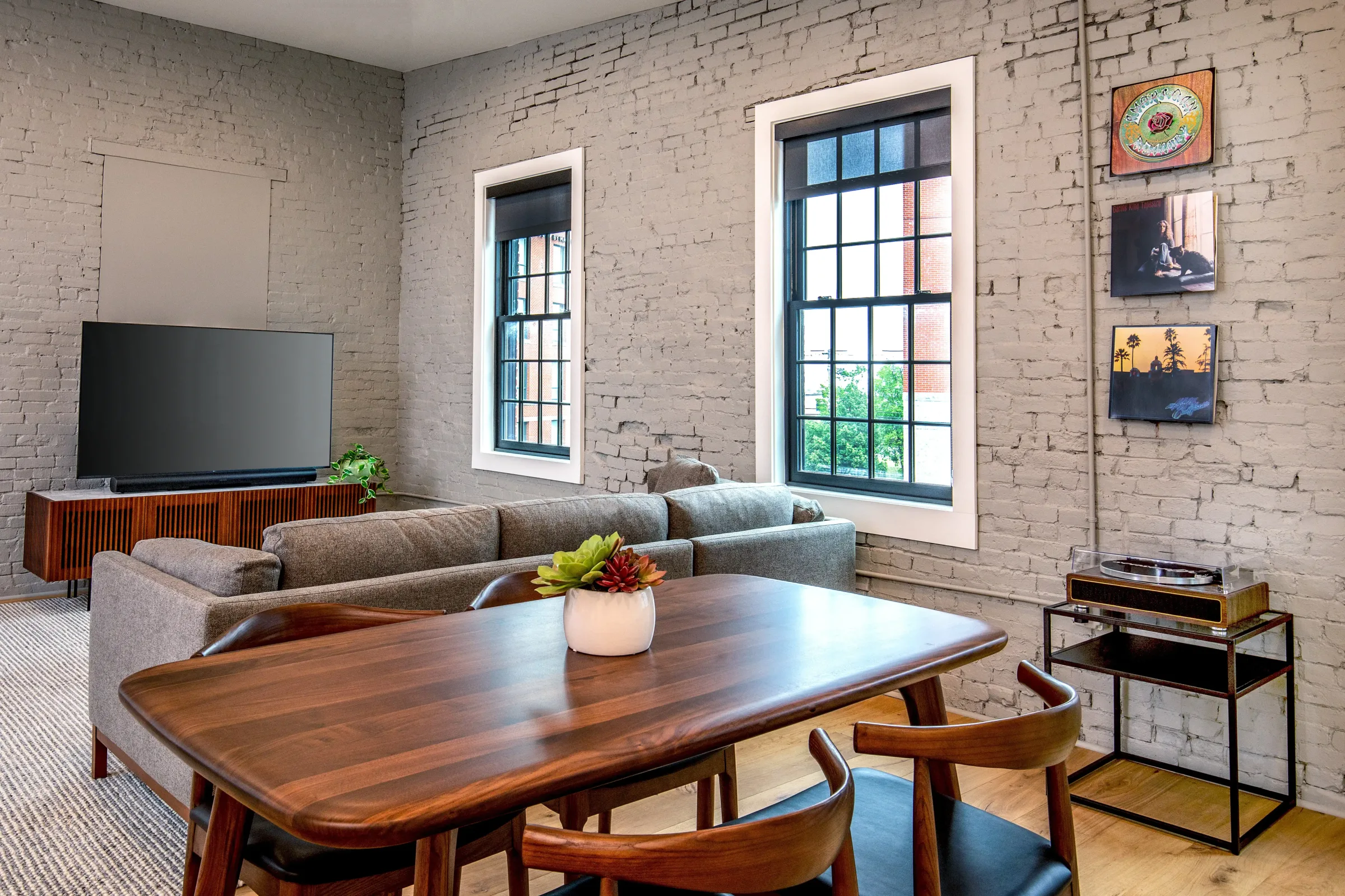 Open-concept living and dining area at the Weldon Hotel, constructed by Woodhull, featuring a gray sectional, wood dining table, record player, and exposed brick walls with framed album art