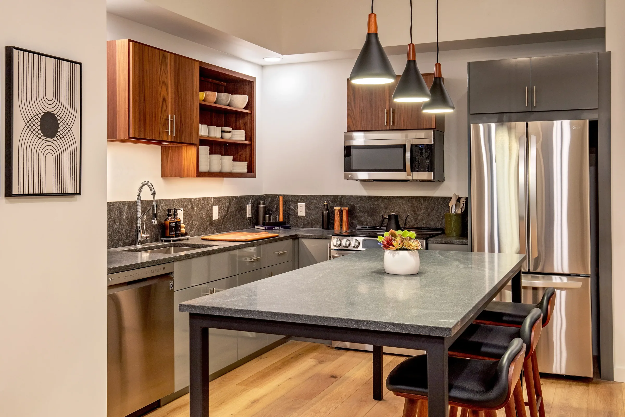 Contemporary kitchen at the Weldon Hotel, constructed by Woodhull, featuring stainless steel appliances, gray and walnut cabinetry, stone countertops, pendant lighting, and a central island with seating