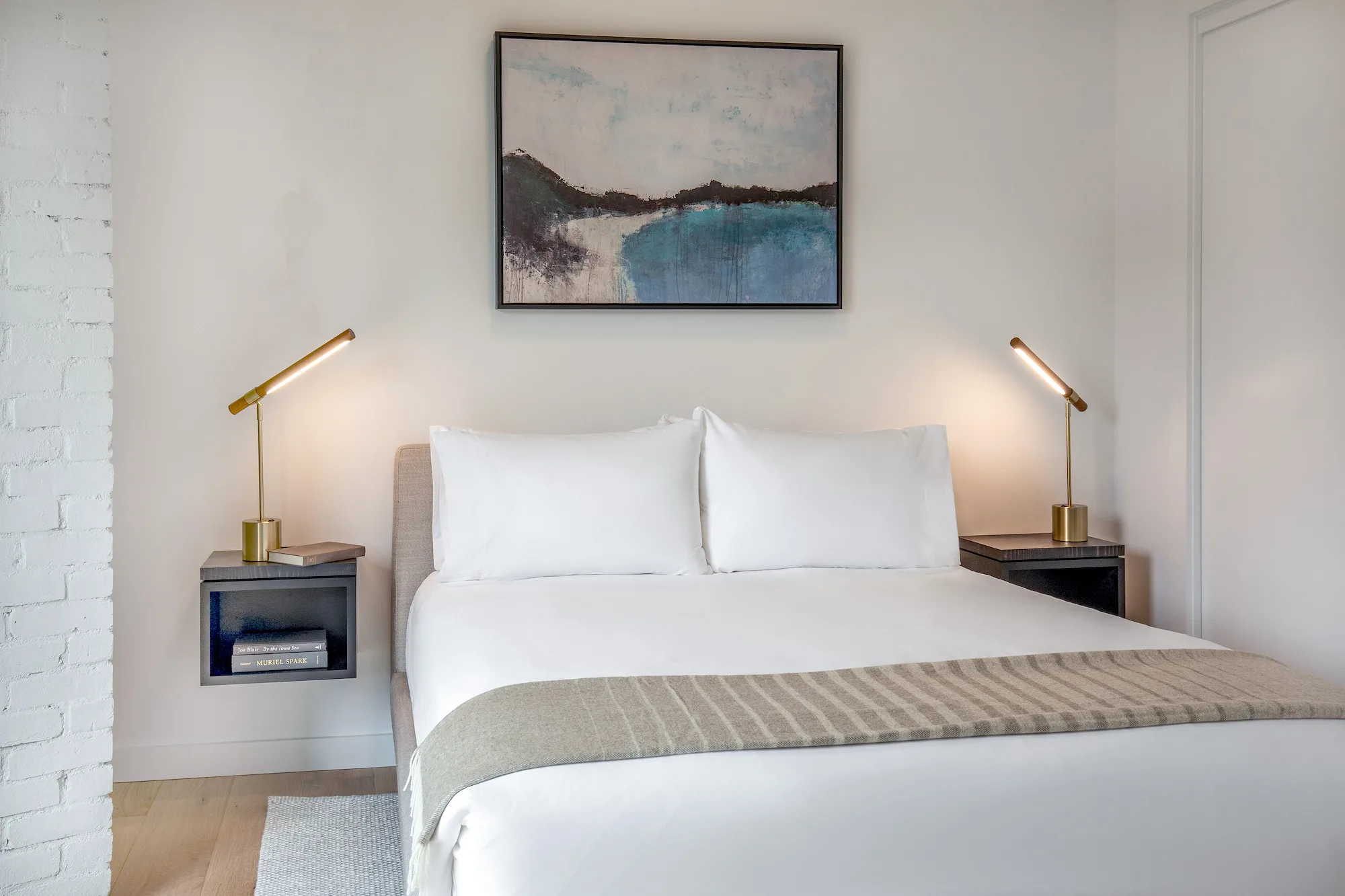 Bright, modern bedroom at the Weldon Hotel, constructed by Woodhull, featuring a queen bed with white linens, brass reading lamps, floating nightstands, and abstract artwork