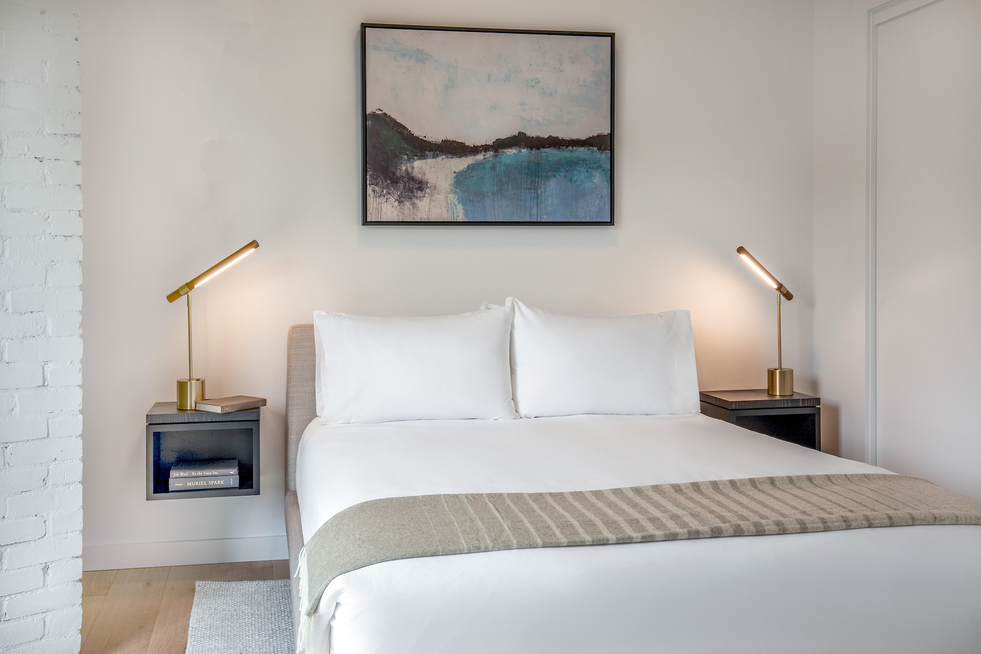Bright, modern bedroom at the Weldon Hotel, constructed by Woodhull, featuring a queen bed with white linens, brass reading lamps, floating nightstands, and abstract artwork