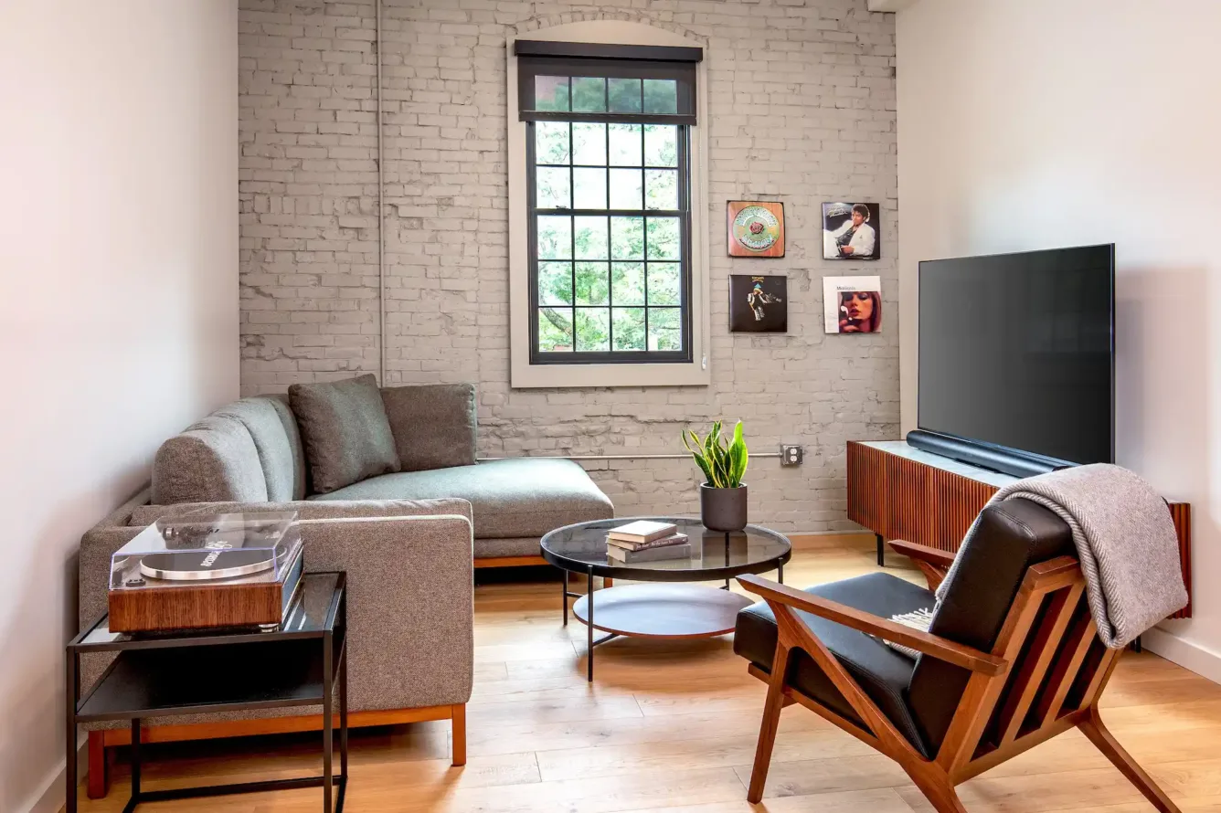 Cozy living space at the Weldon Hotel with a gray sectional sofa, mid-century modern armchair, TV on a wood media console, turntable, coffee table, and album art on a painted brick wall
