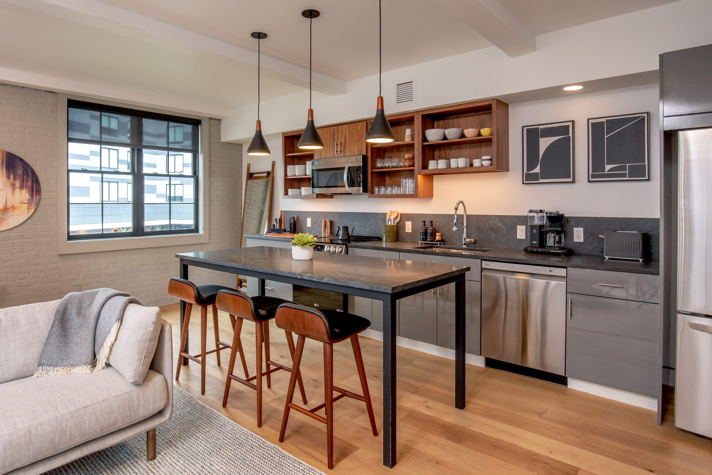 Open-concept kitchen and living area at the Weldon Hotel, constructed by Woodhull, featuring a central island with seating, gray and walnut cabinetry, pendant lighting, stainless steel appliances, and a light-toned sofa