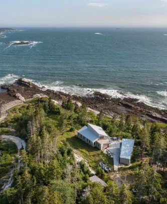 A modern house, designed by a skilled architect, sits on a rocky Maine coastline surrounded by trees and overlooking the ocean under a clear sky.