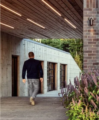 A man in casual clothes walks toward a modern building with wooden and brick elements, designed by a Maine architect, surrounded by plants and trees.