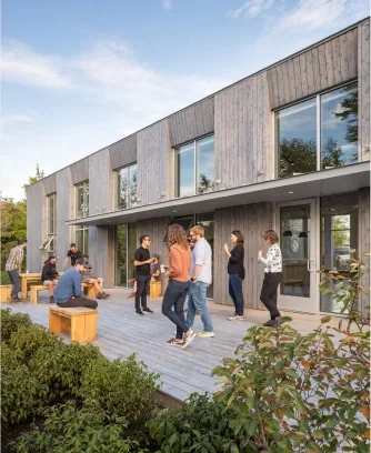 A group socializes on a wooden deck outside a modern building with large windows, showcasing design-build expertise by Maine homebuilders, surrounded by lush greenery.