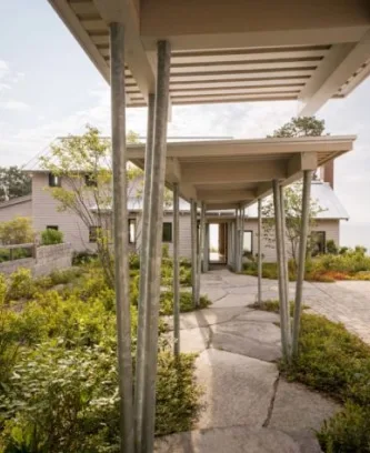 A modern house by Maine homebuilders with a covered walkway supported by metal poles, surrounded by green shrubs and natural stone landscaping.