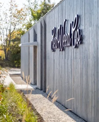 Modern gray wooden building designed by a Maine architect, with a large Bear Weed Co. sign, surrounded by greenery and a gravel path on a sunny day.