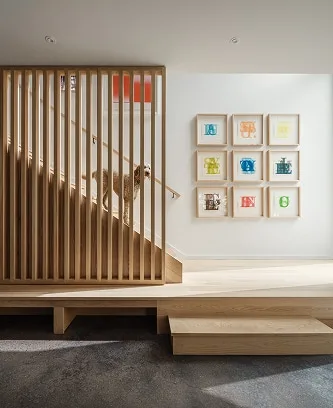 A dog stands on a wooden staircase with vertical slats; nearby, nine colorful framed artworks arranged in a grid showcase the touch of a Maine architect.