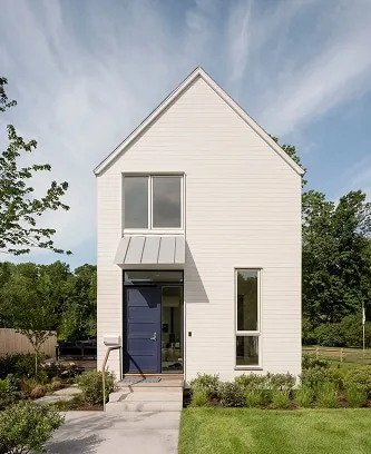 Modern, narrow two-story white house with a blue door, tall windows, and metal awning—crafted by a Maine architect with design-build expertise—nestled among lush greenery.
