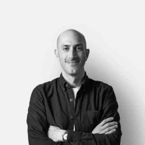 A man with a shaved head, wearing a dark button-up shirt, stands with his arms crossed and smiles at the camera against a plain white background.