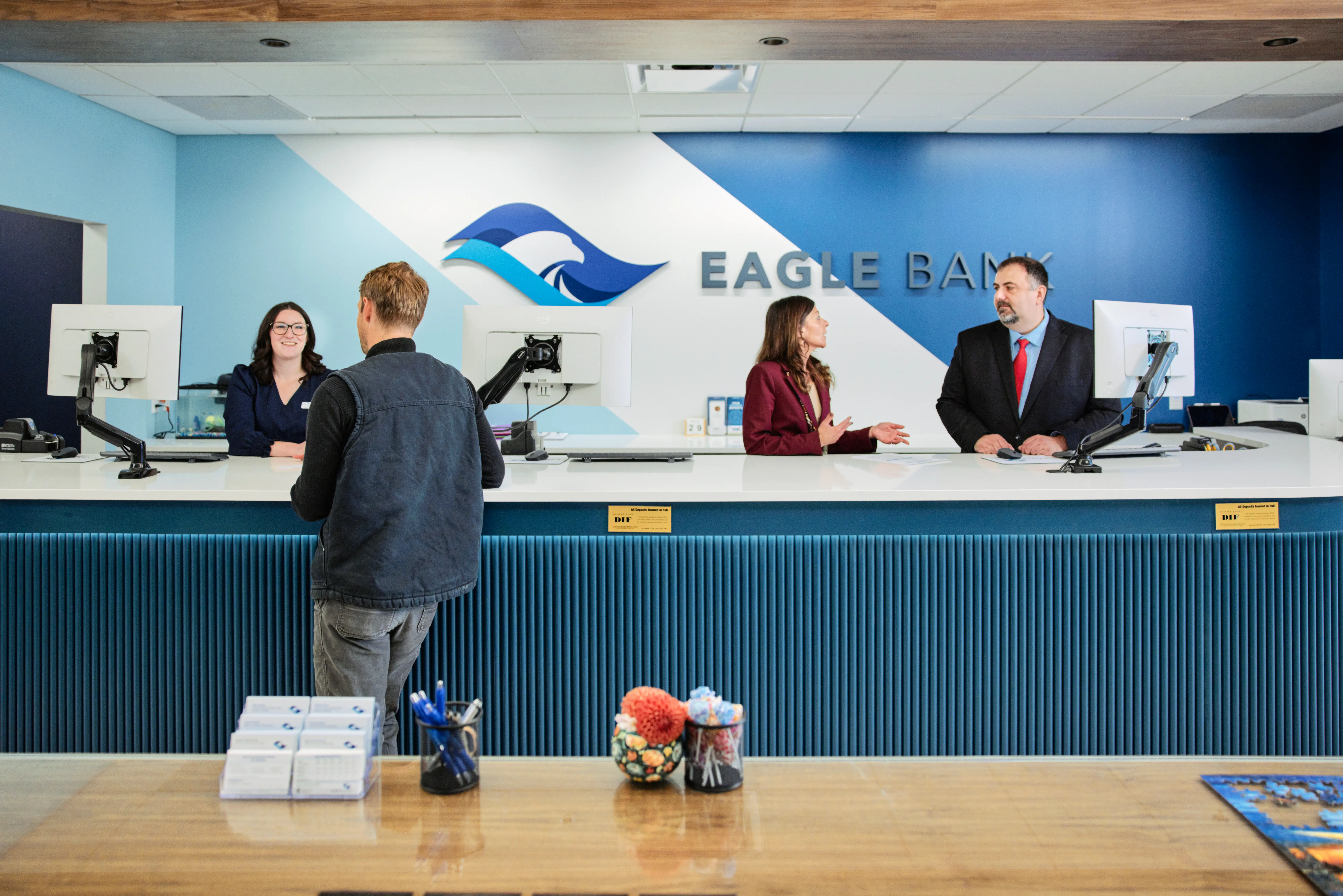 A man stands at a bank counter speaking to a teller, while two other bank employees talk behind the counter. The sign reads Eagle Bank.