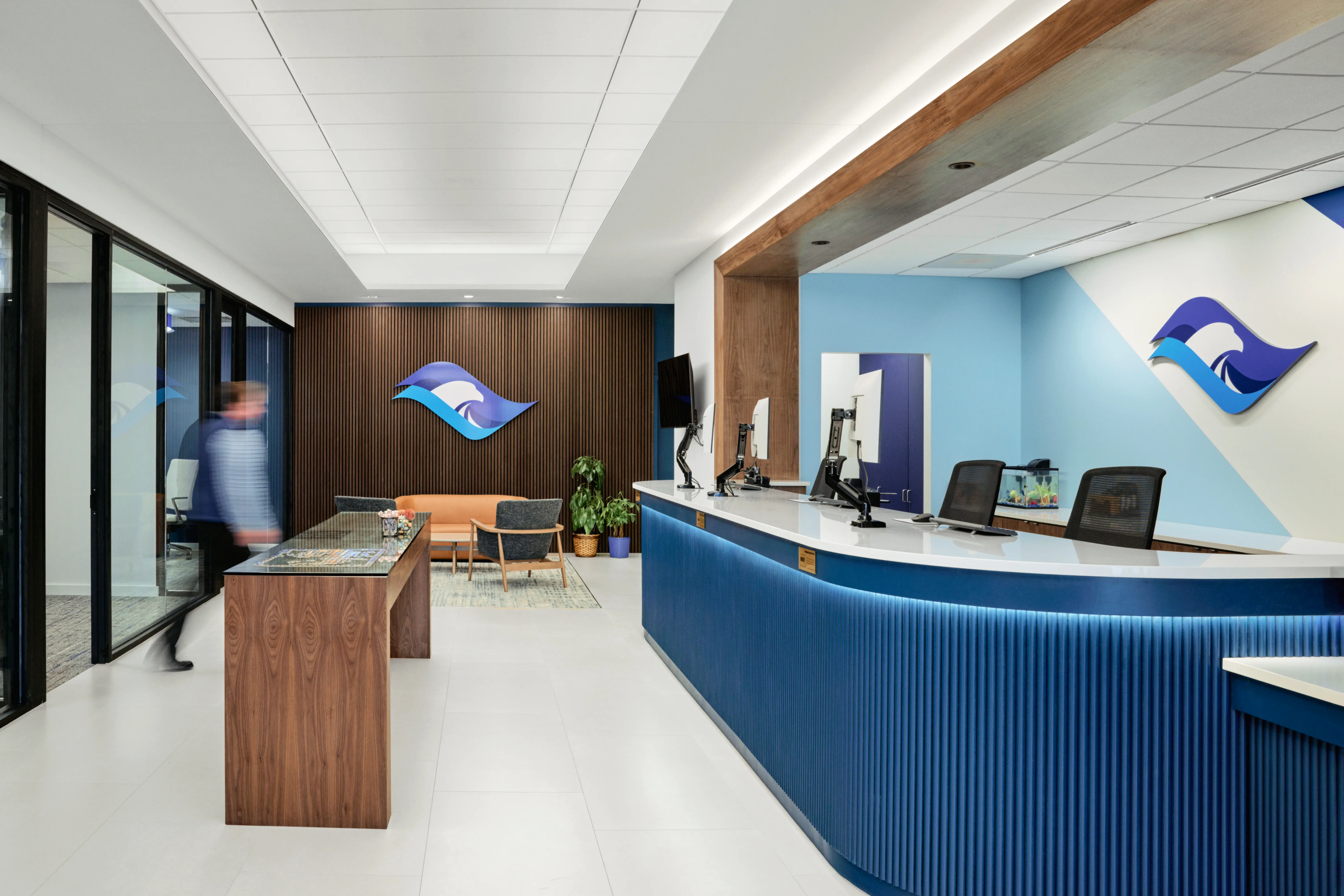 Modern office reception area with a curved blue front desk, wood accents, wall art featuring a wave logo, a small seating area, and a blurred person walking by.