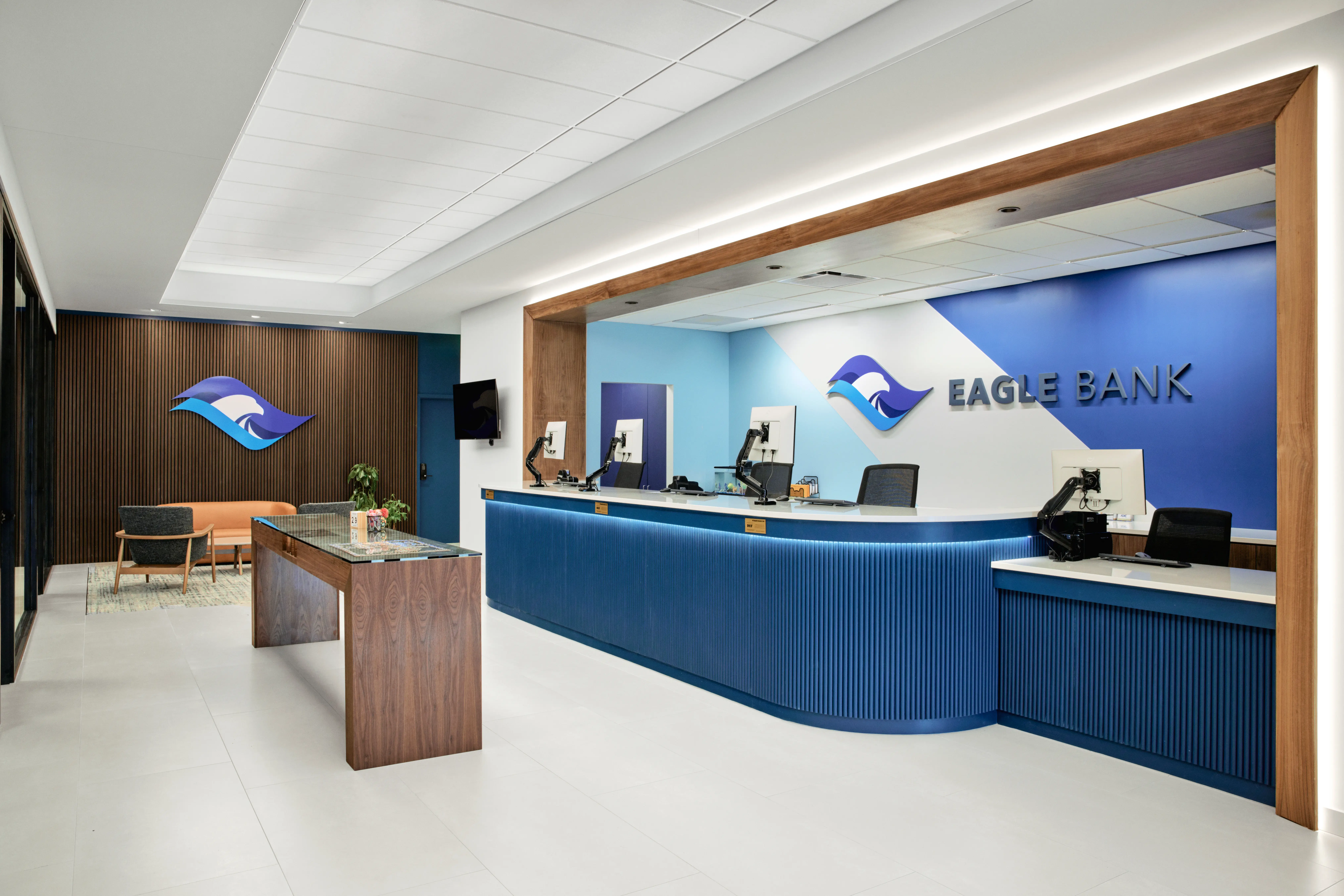 Modern bank interior with a blue and white color scheme, a wood-accented reception desk, and Eagle Bank signage with an eagle logo on the wall behind the counter.