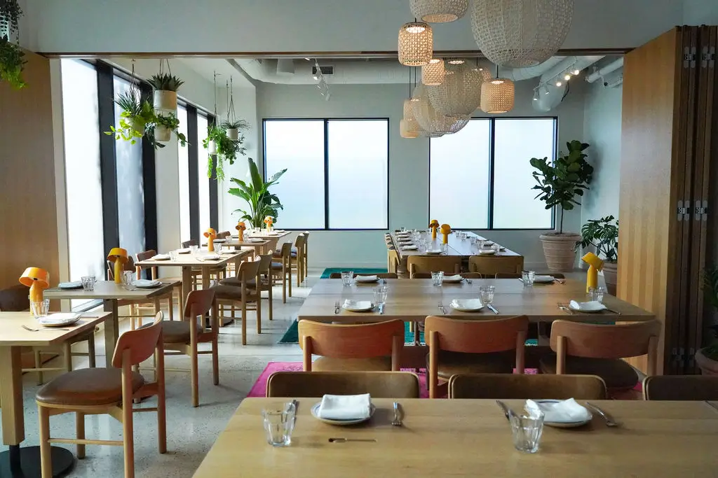 A modern, brightly lit restaurant with wooden tables and chairs, set for dining. Large windows, hanging plants, and decorative lights create a cozy atmosphere.