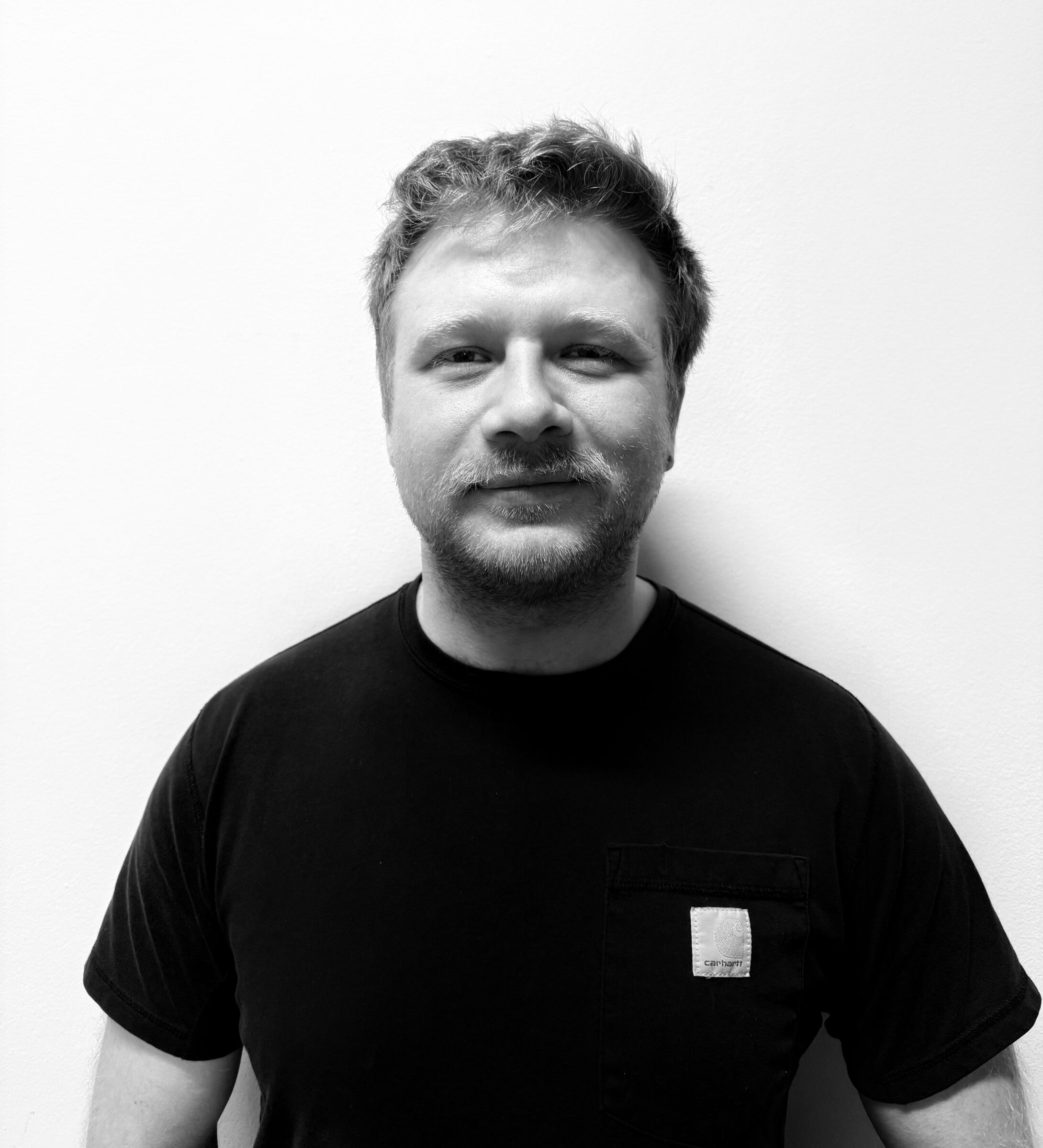 A man with short hair and a beard, wearing a black t-shirt with a pocket, stands against a plain white wall, looking at the camera and smiling slightly. The photo is in black and white.