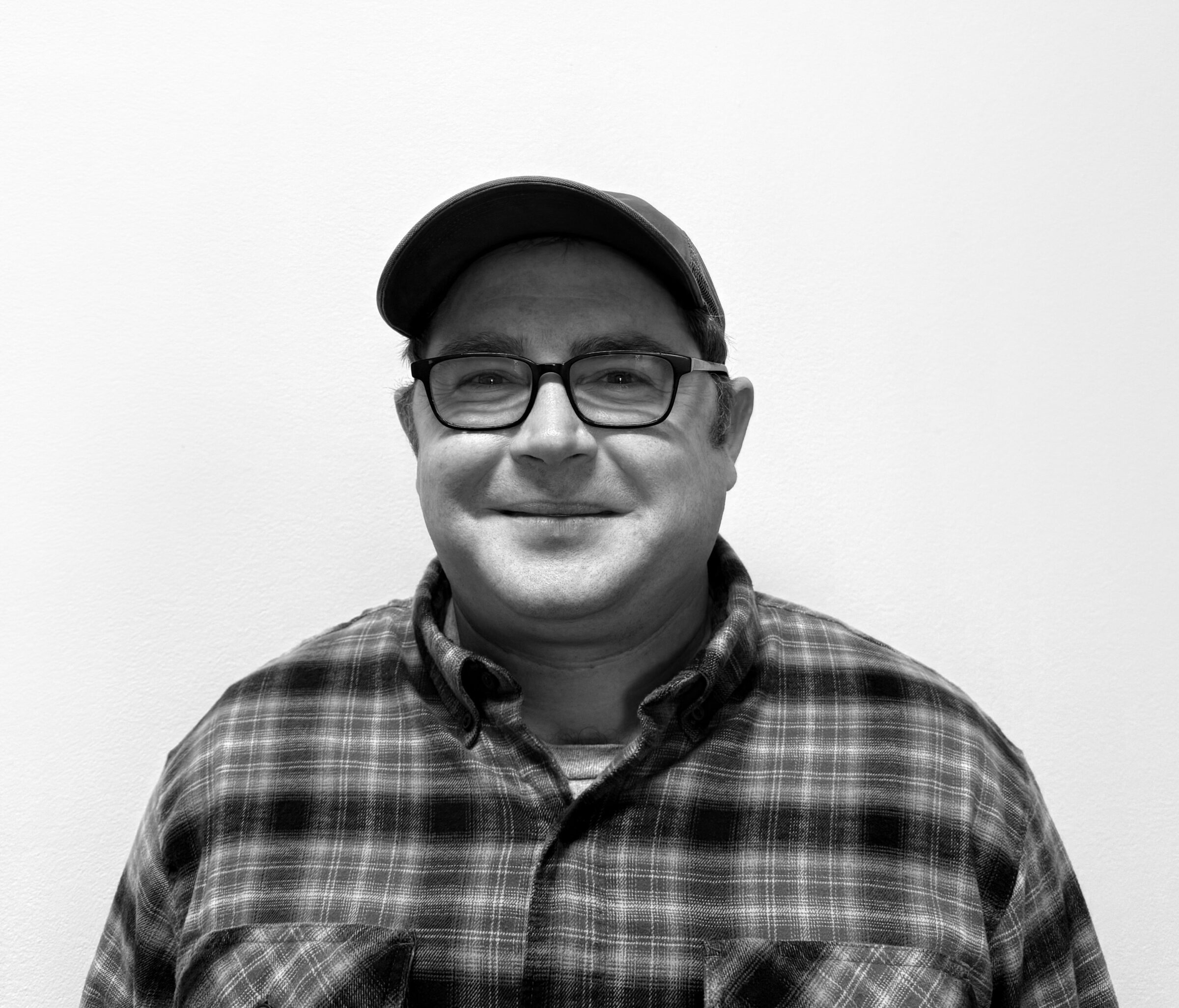 A man wearing glasses, a cap, and a plaid shirt smiles while standing against a plain white background. Black and white photo.