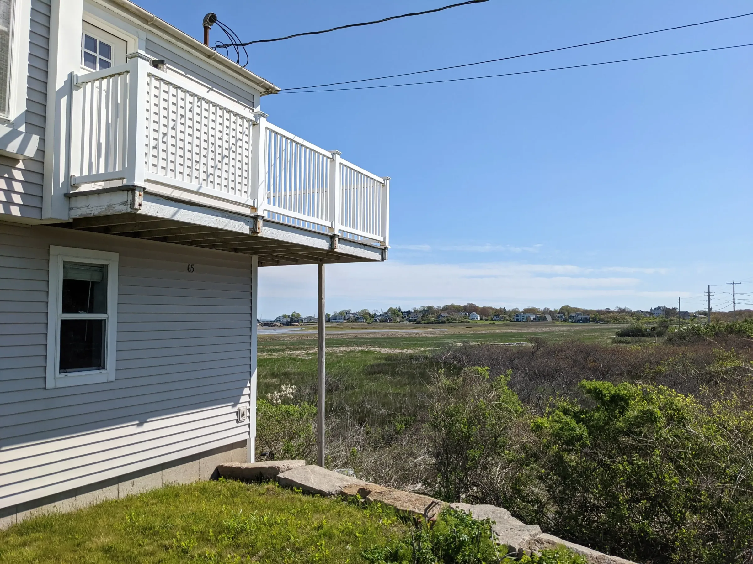 A white house with a balcony overlooks a grassy marshland and distant houses under a clear blue sky.