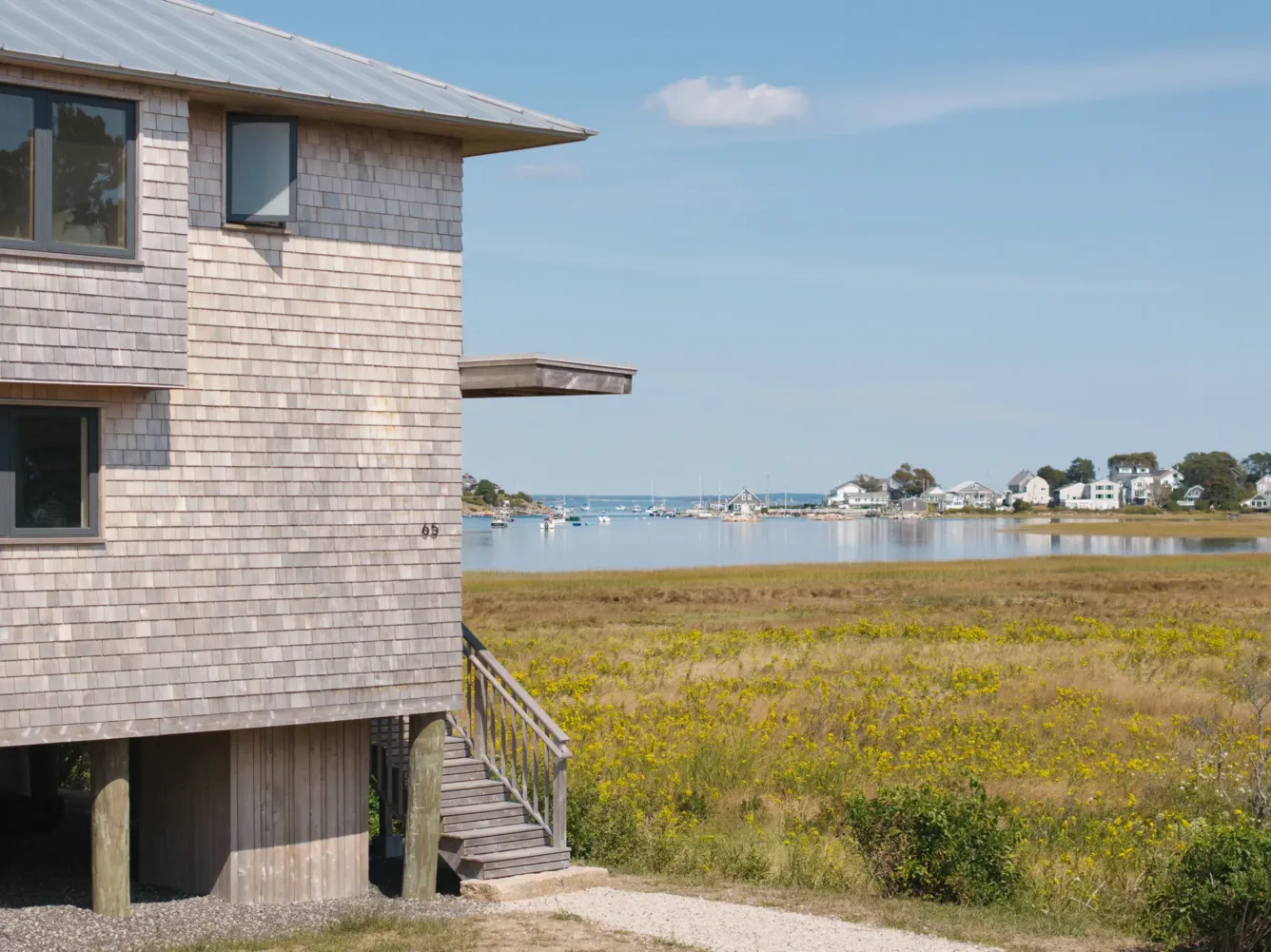 A wooden house with stairs faces a grassy field and a calm bay with boats and houses in the distance under a sunny sky.