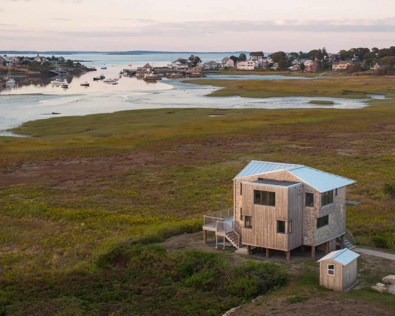 A modern wooden house on stilts stands in a grassy coastal marsh, with water, small boats, and a cluster of houses visible in the background at sunset.
