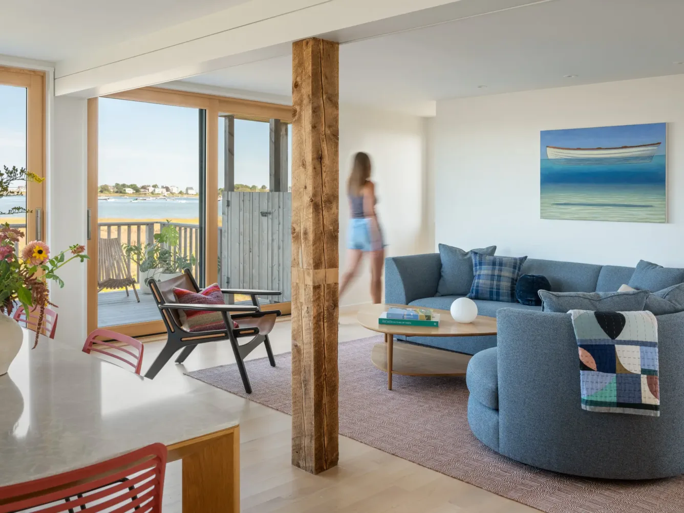 A bright living room with large windows, a round blue sofa, modern chairs, wall art of a boat, and a blurred person walking. A water view is visible outside.