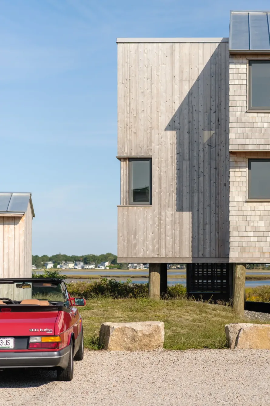 A red convertible car is parked on gravel near a modern wooden house raised on stilts, with water and distant buildings in the background under a clear sky.