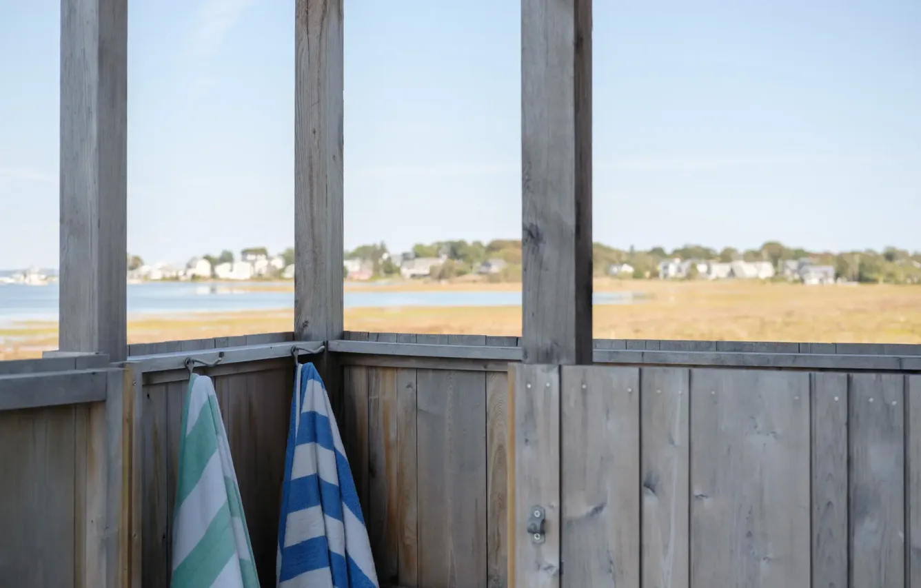 Two striped beach towels hang on a wooden outdoor shower wall, with a scenic view of water, grassy marsh, and distant houses in the background.