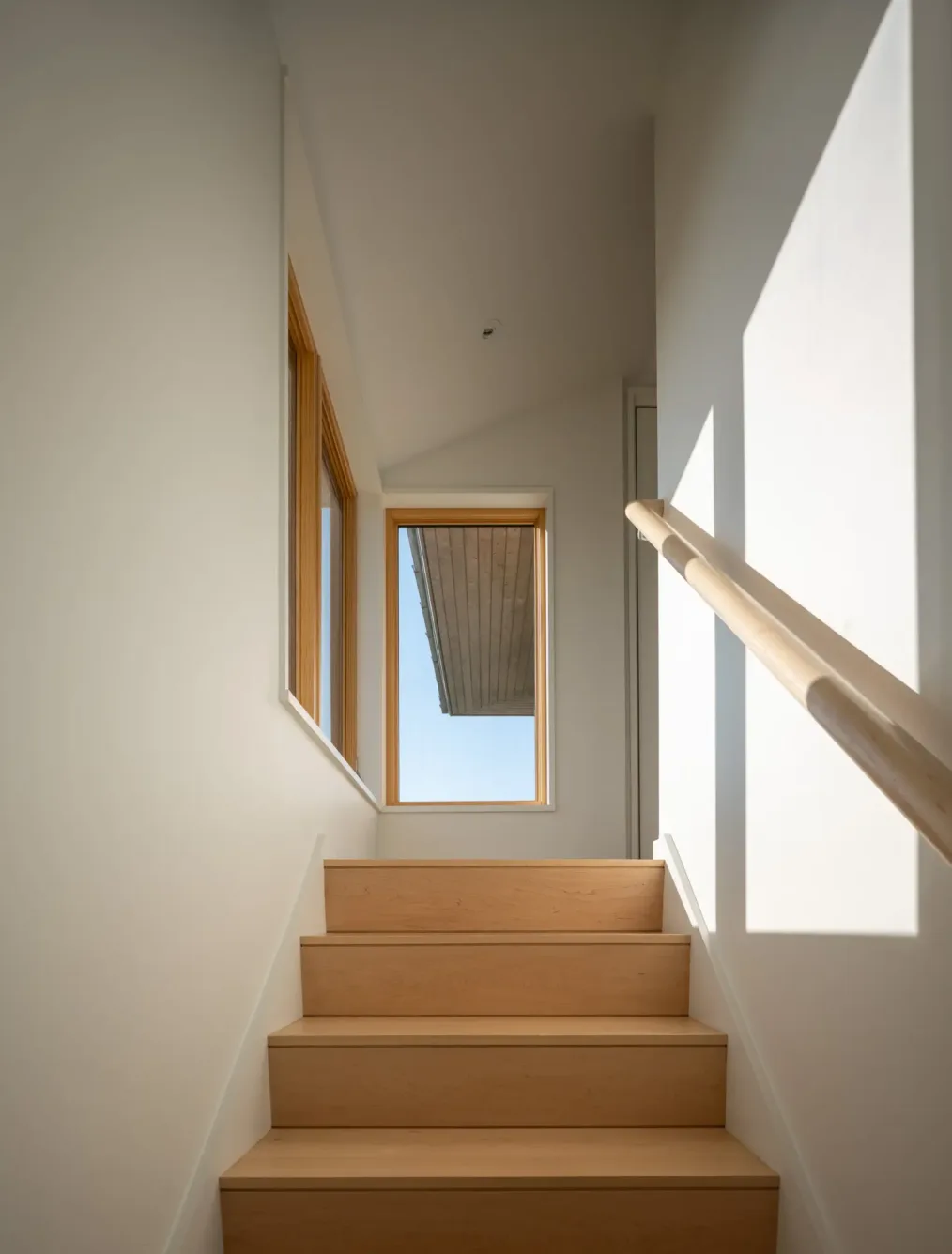 A minimalist wooden staircase leads up to a landing with a large window, letting in sunlight and framing a view of an overhanging roof outside.
