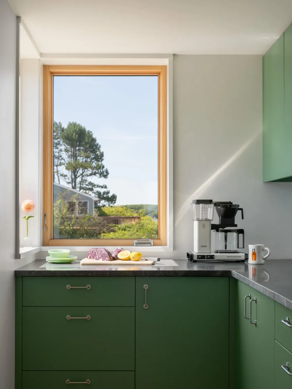 A bright kitchen with green cabinets, a window with a scenic outdoor view, a coffee maker, plates, cups, and sliced bread and lemons on the counter.